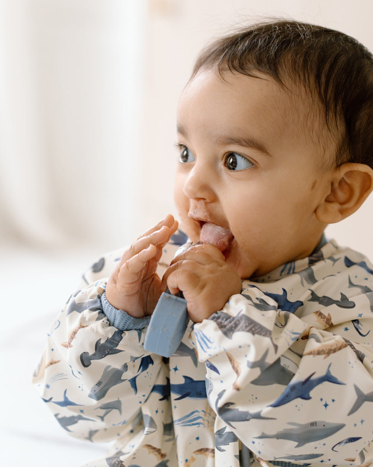 Baby wearing a shark-patterned bib with a blue teether, sitting on a white surface.