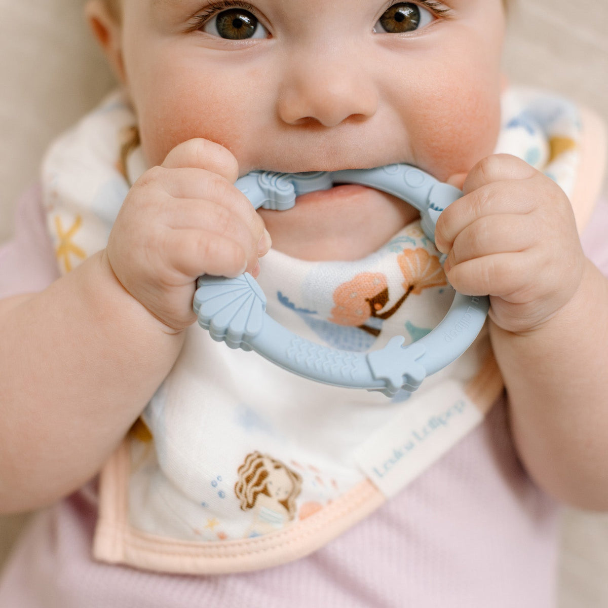 Baby wearing a bib with a teething ring, sitting on a beige surface.