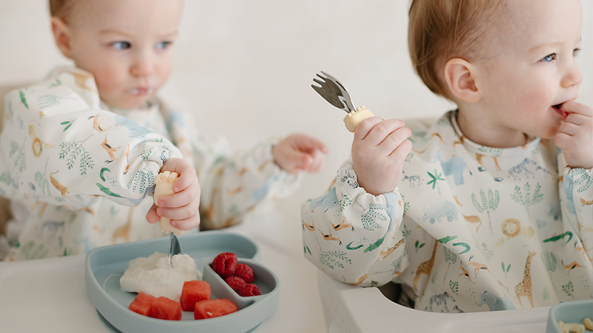 Two toddlers wearing matching long sleeve bibs in the safari jungle print sit at a table, eating from a blue divided tray with a fork and spoon.