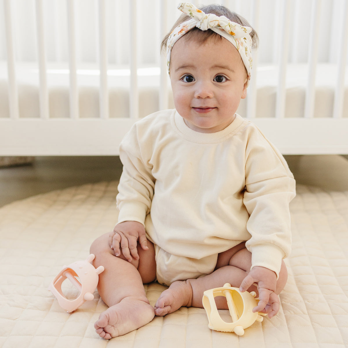 Baby sitting in a crib wearing a white outfit and headband, holding toys.