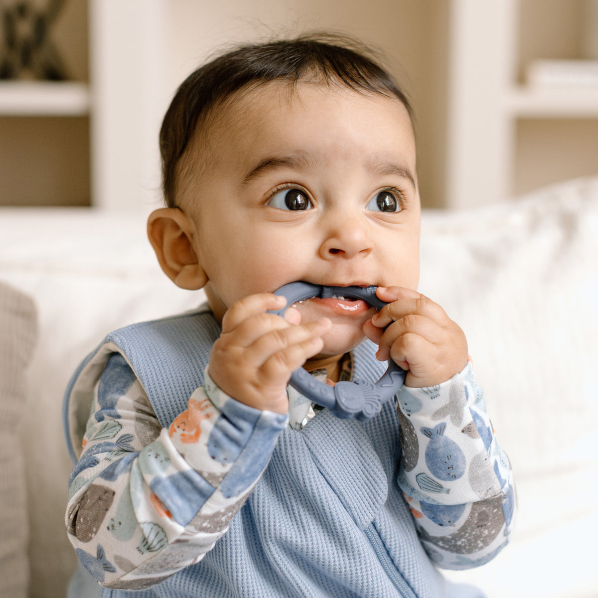 Baby wearing a blue outfit with fish patterns, sitting on a couch and holding a blue object.