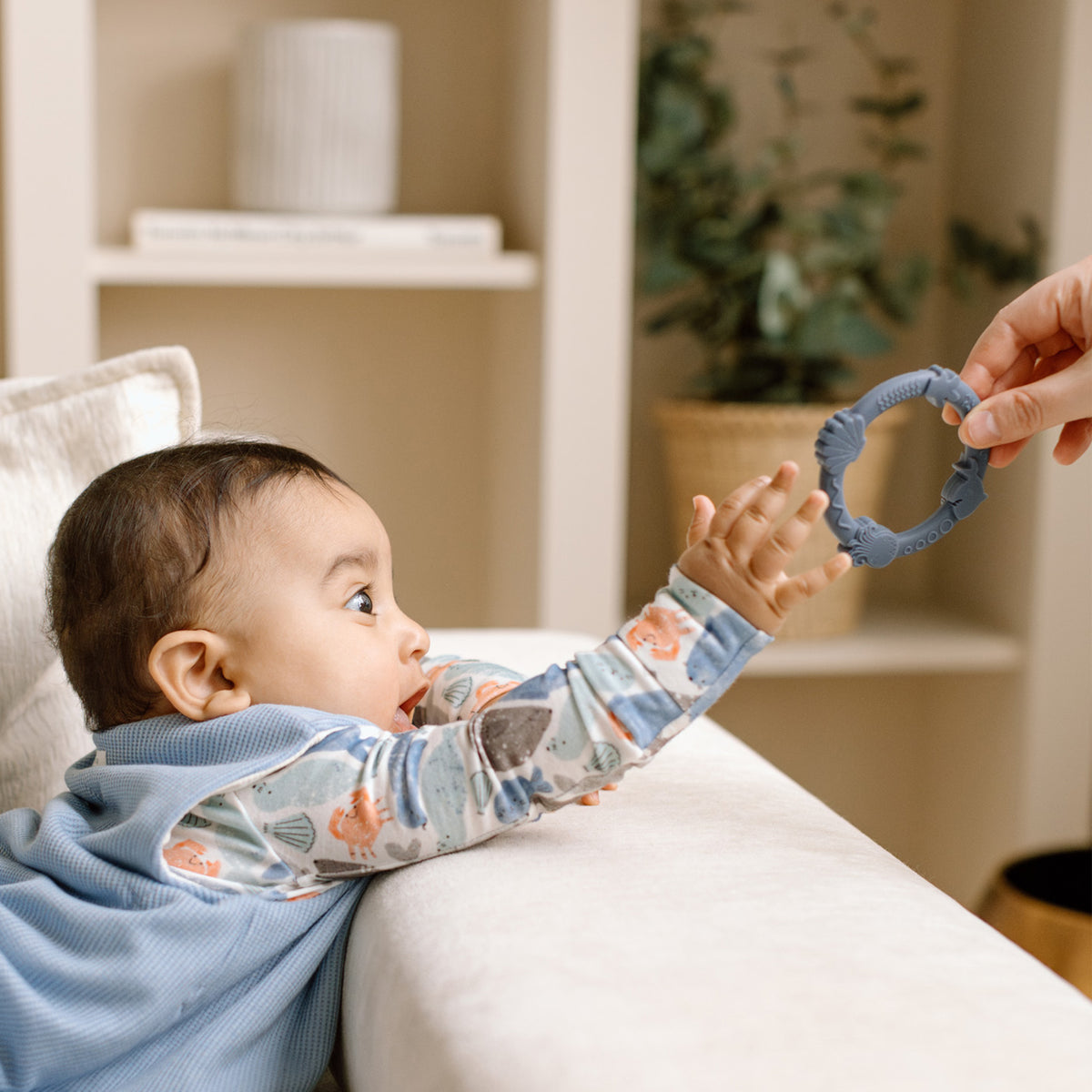 Baby lying on a couch with a teething ring held by an adult hand