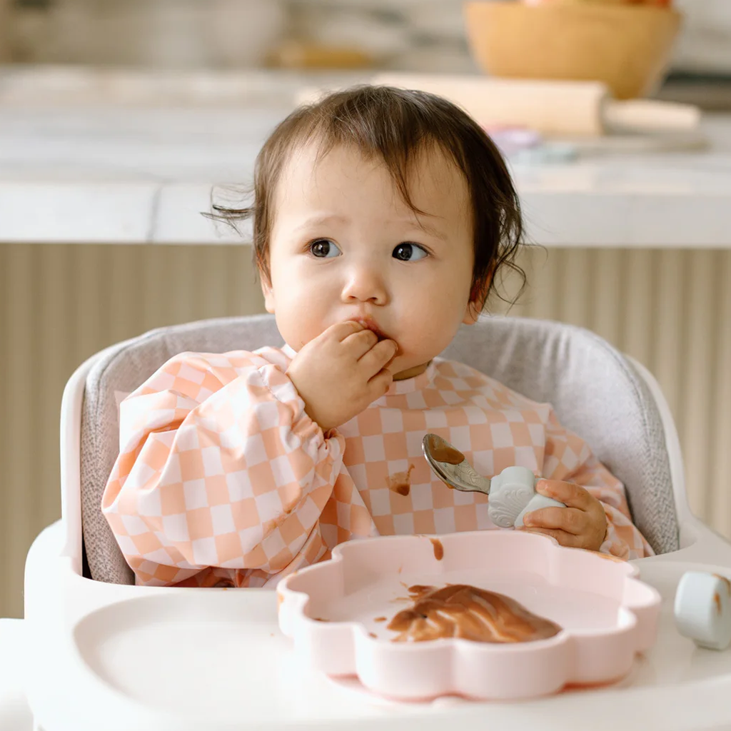 Baby in a high chair eating from a pink bowl with a checkered shirt.