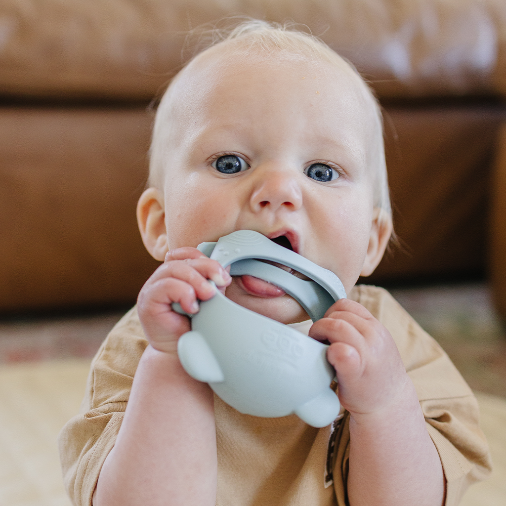 Baby holding a teething toy with a blurred brown couch in the background