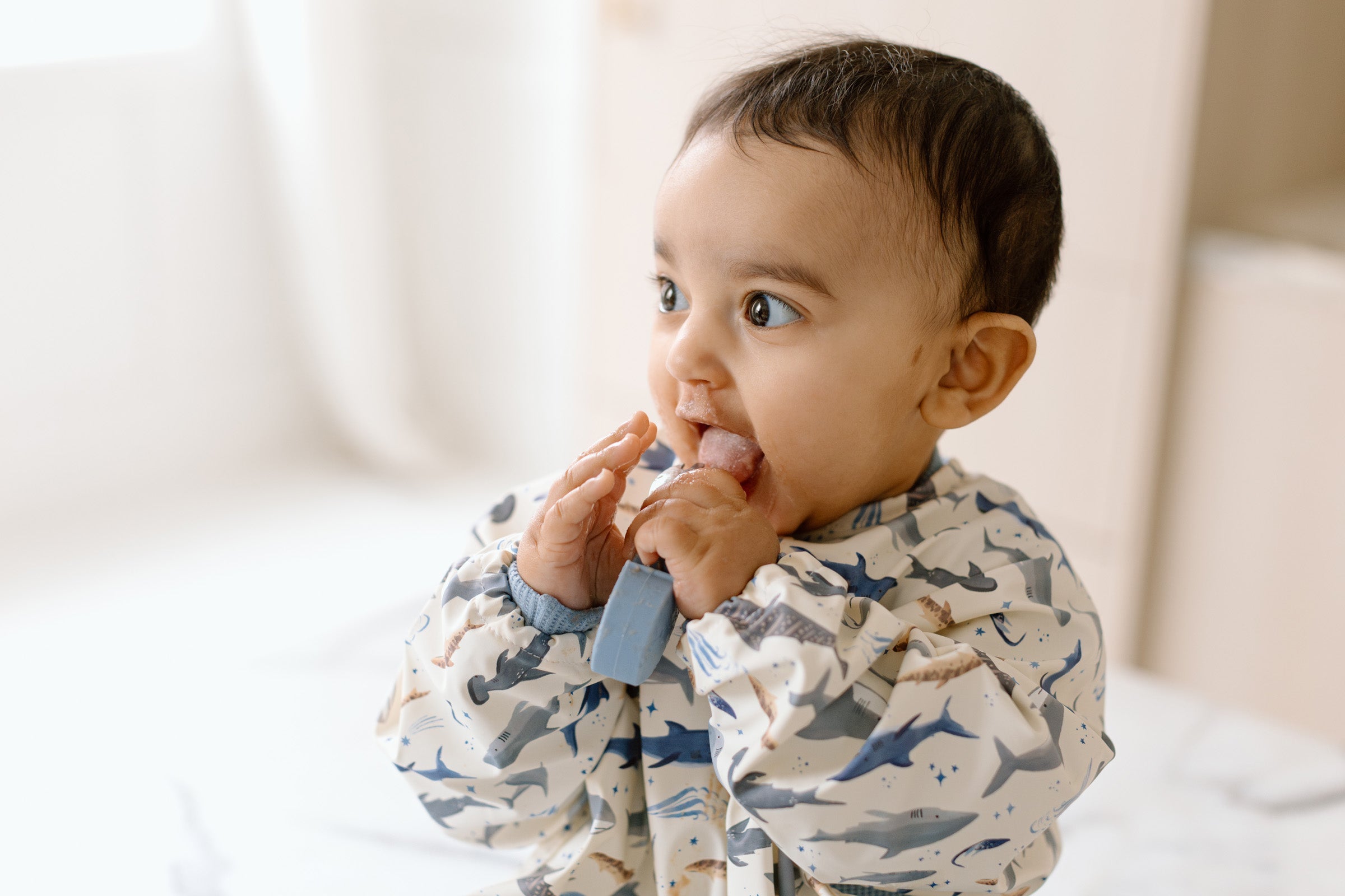 Baby wearing a shark-patterned bib with a blue teether, sitting on a white surface.