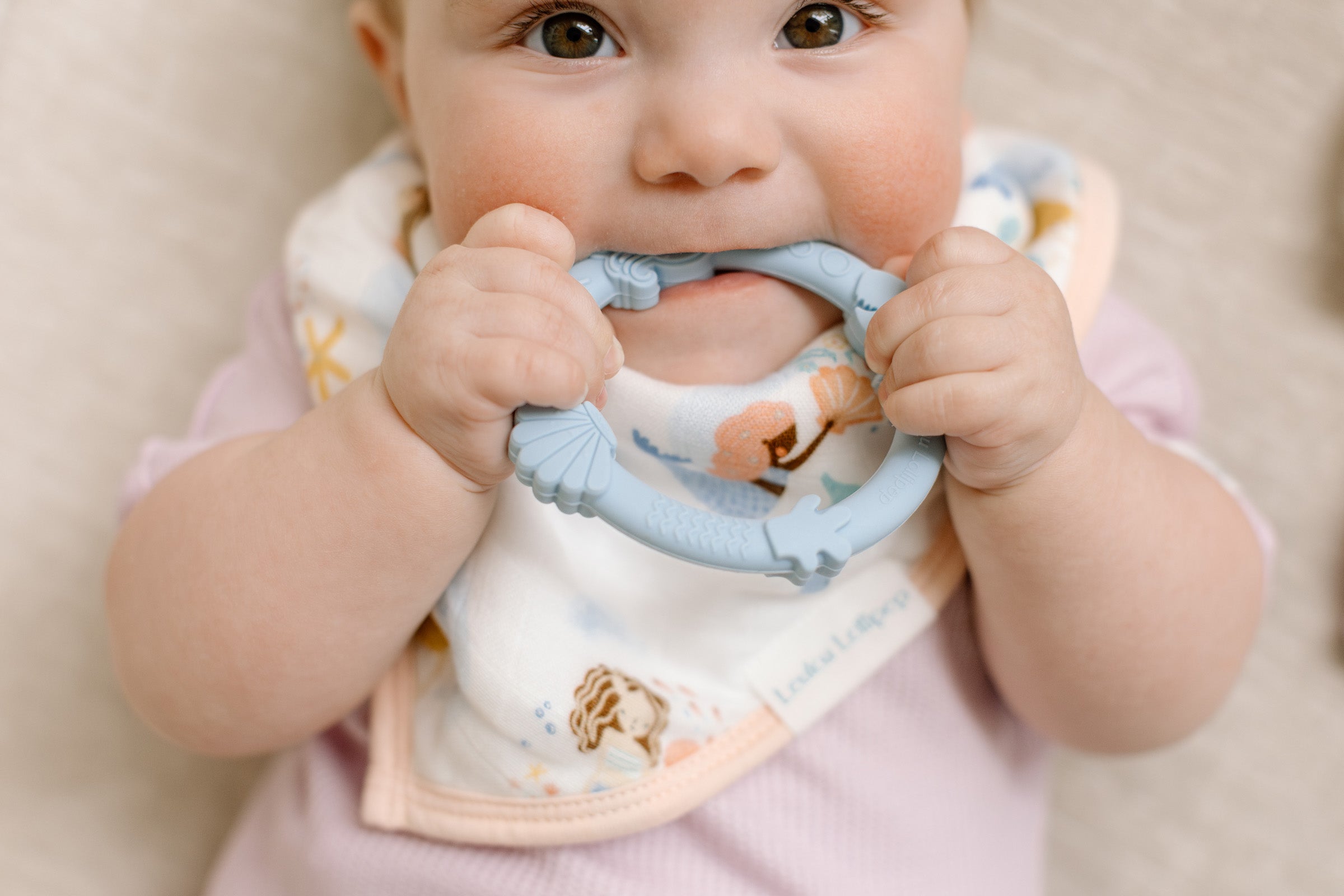 Baby wearing a bib with a teething ring, sitting on a beige surface.